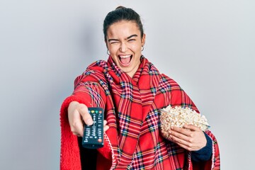 Young caucasian girl wearing blanket eating popcorn using tv control winking looking at the camera...