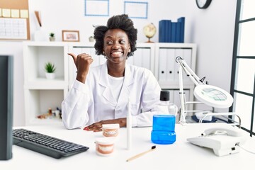 African dentist woman working at medical clinic smiling with happy face looking and pointing to the side with thumb up.