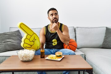 Young hispanic man with beard holding basketball ball cheering tv game serious face thinking about question with hand on chin, thoughtful about confusing idea