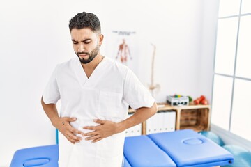 Young handsome man with beard working at pain recovery clinic with hand on stomach because indigestion, painful illness feeling unwell. ache concept.