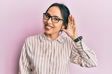 Young hispanic girl wearing casual clothes and glasses smiling with hand over ear listening and hearing to rumor or gossip. deafness concept.