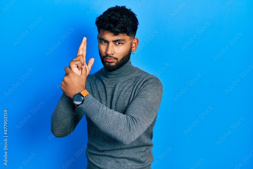 Wall mural Arab man with beard wearing turtleneck sweater holding symbolic gun with hand gesture, playing killing shooting weapons, angry face