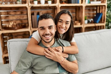 Young latin couple sitting on the sofa hugging at home. - obrazy, fototapety, plakaty