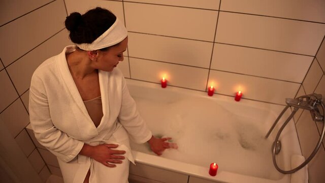 Overhead View Of A Charming Serene Woman In White Waffle Bathrobe Sitting On The Edge Of A White Bath Tub And Filling Up Water For Taking Relaxing Foam Bath. Body Care, Hygiene And Purity Concept