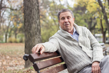 Portrait of a pensive senior man sitting on the bench, in the public park, outdoors. Old man relaxing outdoors and looking at camera. Portrait of elderly man enjoying retirement