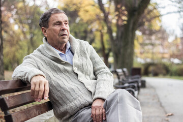 Fototapeta premium Portrait of a pensive senior man sitting on the bench, in the public park, outdoors. Old man relaxing outdoors and looking away. Portrait of elderly man enjoying retirement