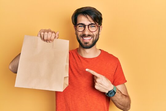 Young hispanic man holding paper bag with bread smiling happy pointing with hand and finger