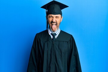 Middle age hispanic man wearing graduation cap and ceremony robe sticking tongue out happy with funny expression. emotion concept.