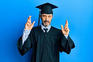 Middle age hispanic man wearing graduation cap and ceremony robe gesturing finger crossed smiling with hope and eyes closed. luck and superstitious concept.