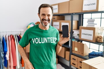 Middle age man with beard wearing volunteer t shirt holding smartphone winking looking at the...