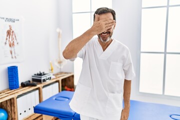 Middle age man with beard working at pain recovery clinic smiling and laughing with hand on face covering eyes for surprise. blind concept.