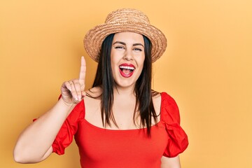 Young hispanic woman wearing summer hat showing and pointing up with finger number one while smiling confident and happy.