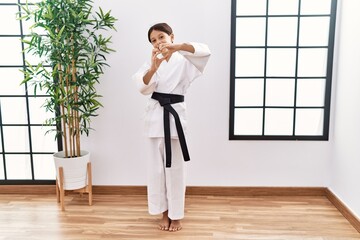 Young hispanic girl wearing karate kimono and black belt smiling in love doing heart symbol shape with hands. romantic concept. © Krakenimages.com