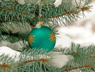 Fototapeta na wymiar Green Christmas ball hanging on a snow-covered branch of a Christmas tree on a festive background of white snow and snowy bokeh with copy space.