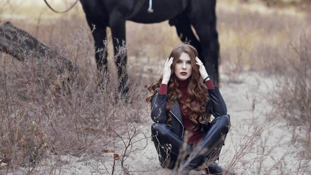beautiful girl sida on the ground with her legs crossed posing on the background of a black horse
