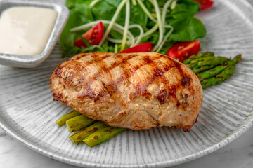 Whole grilled chicken breast, asparagus and tomato salad with arugula in a festive plate on a marble background. Restaurant banquet menu.