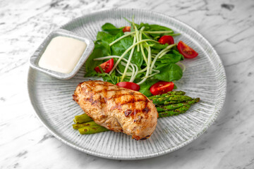 Whole grilled chicken breast, asparagus and tomato salad with arugula in a festive plate on a marble background. Restaurant banquet menu.