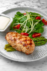 Whole grilled chicken breast, asparagus and tomato salad with arugula in a festive plate on a marble background. Restaurant banquet menu.