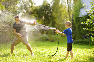 Funny little boy with his father playing with garden hose in sunny backyard. Preschooler child having fun with spray of water. Summer outdoors activity for kids.