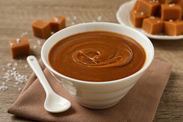 Yummy salted caramel in bowl and candies on wooden table, closeup