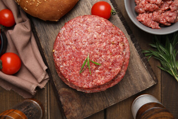 Raw hamburger patties with rosemary and tomatoes on wooden table, flat lay