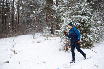 Skier with a backpack and hat with pompom with ski poles in his hands on background of a snowy forest. Cross-country skiing in winter forest, outdoor sports, healthy lifestyle, winter sports tourism.