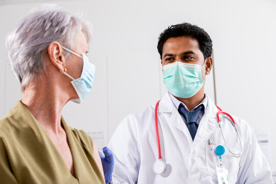 A Young Indian Male Medical Doctor Consults with an Elderly Senior Female Patient about Covid-19 Vaccine Injection Wearing Generic ID Badge, Lab Coat, Gloves and Mask in Hospital or Health Clinic.