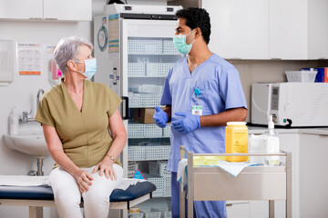 A Young Indian Male Medical Nurse Consults with an Elderly Senior Female Patient about Covid-19 Vaccine Injection Wearing Generic ID Badge, Scrubs, Gloves and Mask in Hospital or Health Clinic.
