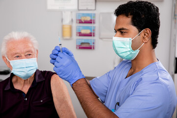 A Young Indian Male Medical Nurse Administering a Covid-19 Vaccine with a Syringe Needle to an Elderly Senior Male Patient Wearing Generic ID Badge, Gloves and Mask in Hospital or Health Clinic.