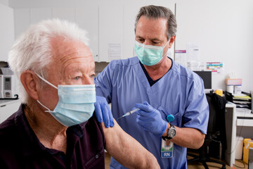 Fototapeta na wymiar A White Male Medical Nurse Administering a Covid-19 Vaccine with a Syringe Needle to an Elderly Senior Male Patient Wearing Generic ID Badge, Gloves, Scrubs and Mask in Hospital or Health Clinic.