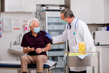 A White Male Medical Doctor Consults with an Elderly Senior Male Patient about Covid-19 Vaccine Injection Wearing Generic ID Badge, Lab Coat, Gloves and Mask in Hospital or Health Clinic.