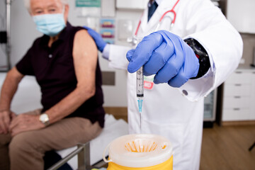 A Young Indian Male Medical Doctor Disposes of Syringe Needle Safely in Sharps Collector after a Covid-19 Vaccine Injection Wearing Gloves, Mask and Generic ID Badge in Hospital or Health Clinic. 
