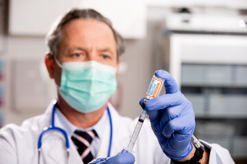 A White Male Medical Doctor Extracting a Covid-19 Vaccine with a Syringe Needle Wearing Generic ID Badge, Gloves, Surgical Protective Mask, Lab Coat and Stethoscope in Hospital or Health Clinic.