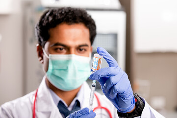 A Young Indian Male Medical Doctor Extracting a Covid-19 Vaccine Injection with a Syringe Needle Wearing Generic ID, Gloves, Mask, White Lab Coat and Stethoscope in Hospital or Health Clinic.