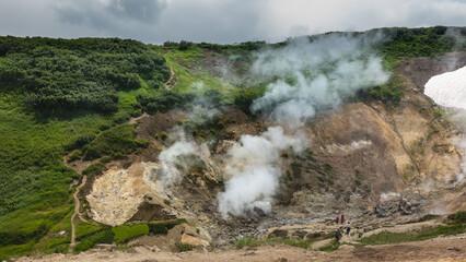 Columns of steam from hot springs rise in the geothermal valley.  The path winds along the mountainside, among the green vegetation. Tiny silhouettes of people are visible. Kamchatka