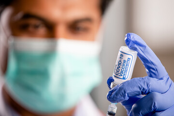 A Young Indian Male Medical Doctor Extracting a Covid-19 Vaccine Injection with a Syringe Needle Wearing Generic ID, Gloves, Mask, White Lab Coat and Stethoscope in Hospital or Health Clinic.