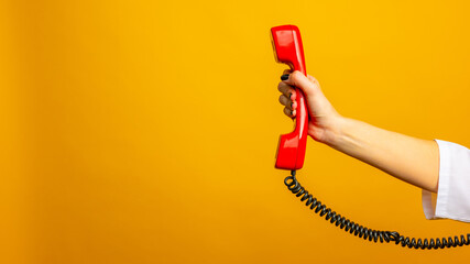 Closeup view of woman holding red corded telephone handset on yellow background, space for text. Hotline concept - obrazy, fototapety, plakaty