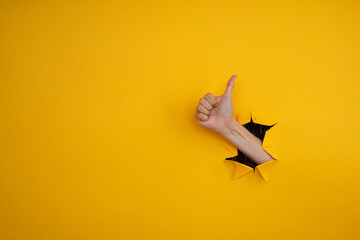 Hand showing a thumb up sign through a ripped hole in yellow paper wall. Well done, good job...