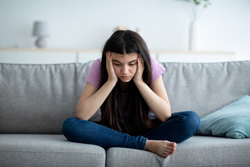 Sad Indian teenage girl sitting on sofa at home with head in hands, suffering from lack of...