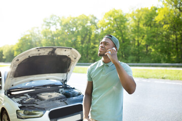 Stressed black guy standing near broken vehice, having mobile phone conversation with breakdown road service outdoors