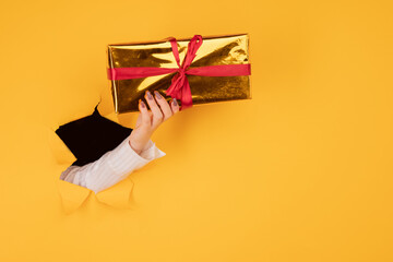 A female hand in a torn hole on a yellow background holds a golden box with a gift. Hand through torn paper