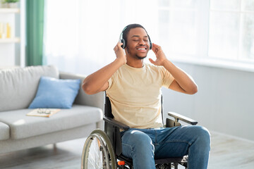 Joyful impaired black guy in wheelchair listening to music, using headphones at home, copy space