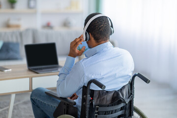 Back view of paraplegic black guy in wheelchair wearing headphones, using laptop with mockup for design at home