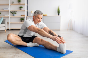 Fototapeta Home workout. Sporty mature man stretching her leg on yoga mat in living room, free space obraz