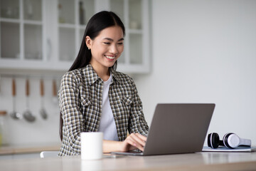 Positive asian lady freelancer sitting at kicthen, working online