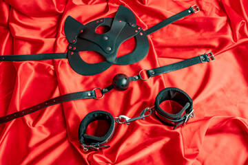 Adult sex games. BDSM items. Leather mask cat, gag ball, handcuffs on a red satin sheet. - obrazy, fototapety, plakaty