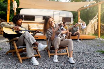 Young black guy playing guitar and singing, Caucasian lady holding dog near RV, camping with...