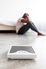 Fototapeta premium Depressed plus size black lady sitting on floor at home, disappointed in her slimming result, selective focus on scales