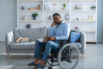 Impaired black man in wheelchair reading notebook, studying or working from home, full length