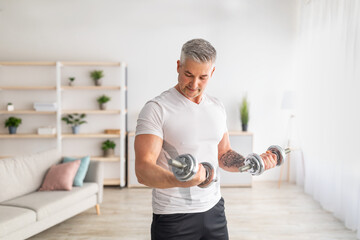 Athletic mature man doing dumbbell workout at home, working on arms strength, looking at his...